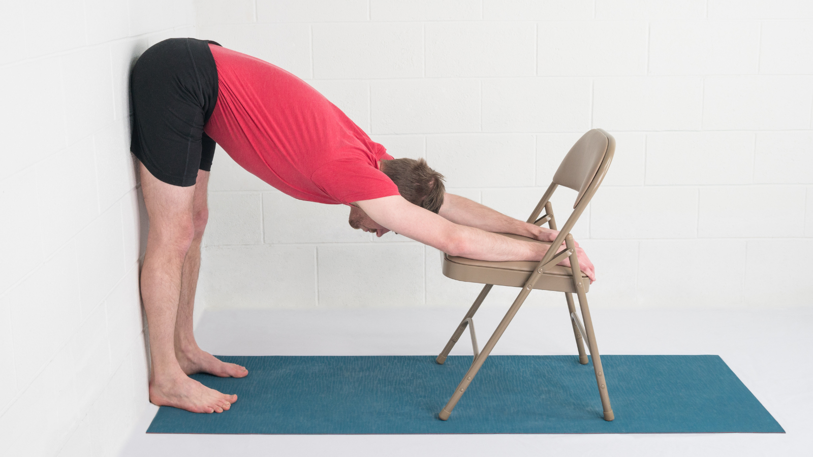 Man practicing Uttanasana (Standing Forward Bend Pose) variation with chair at the wall