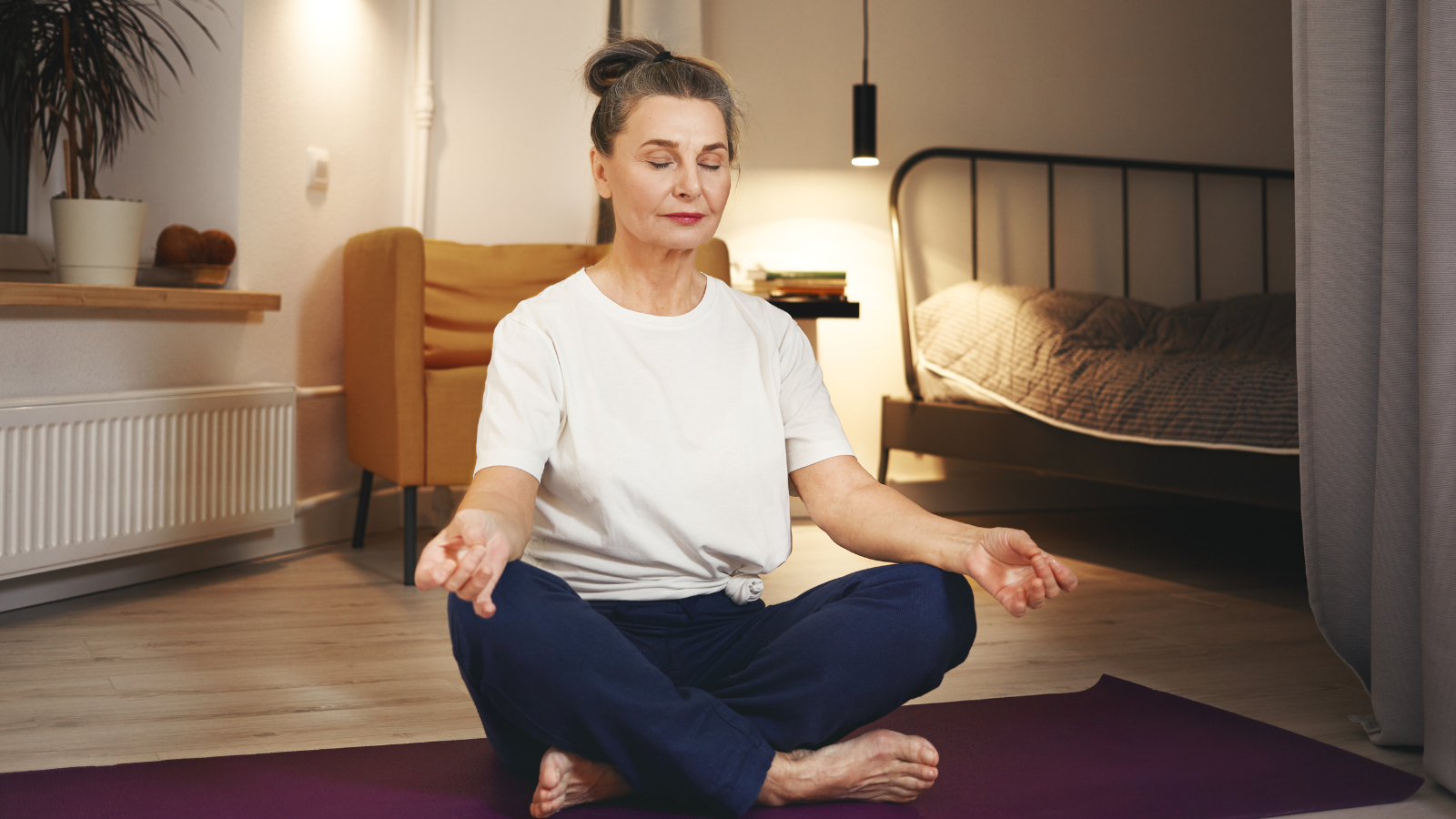 Senior woman sitting barefooted on floor at home, keeping legs crossed on mat, doing meditation after yoga practice, having calm peaceful facial expression