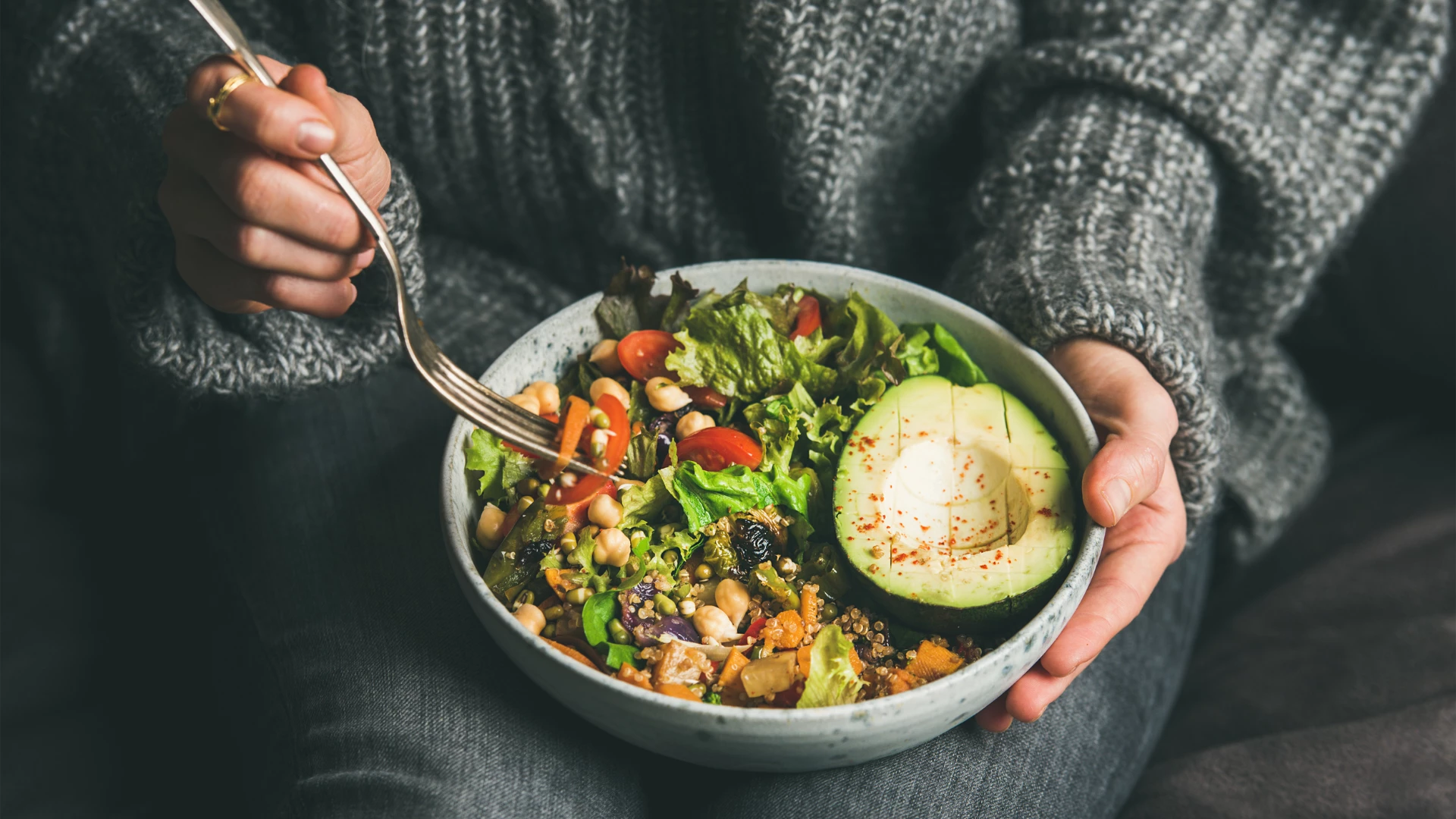 Healthy vegetarian dinner. Woman in jeans and warm sweater holding bowl with fresh salad, avocado, grains, beans, roasted vegetables, close-up. Superfood, clean eating, vegan, dieting food concept