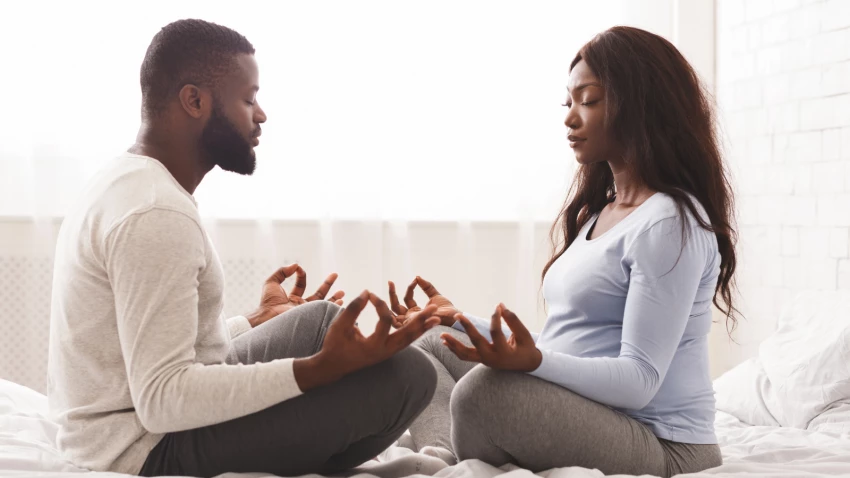 Pregnant woman doing yoga with her husband at home, sitting on bed face to face with closed eyes