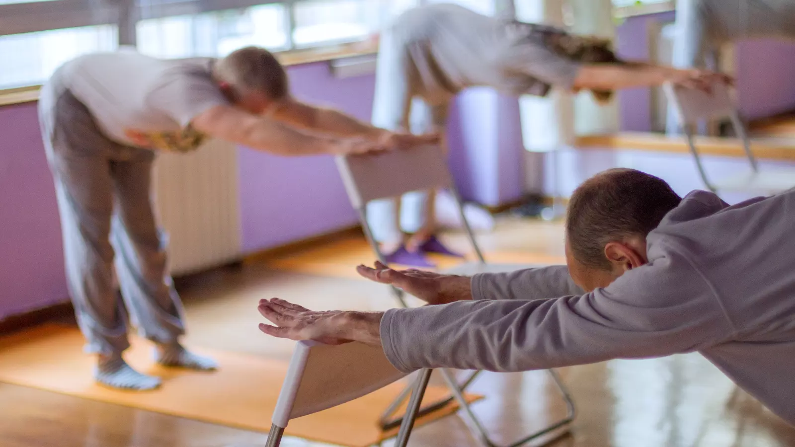 A group pf people using the back of a chair to help them do a yoga pose.