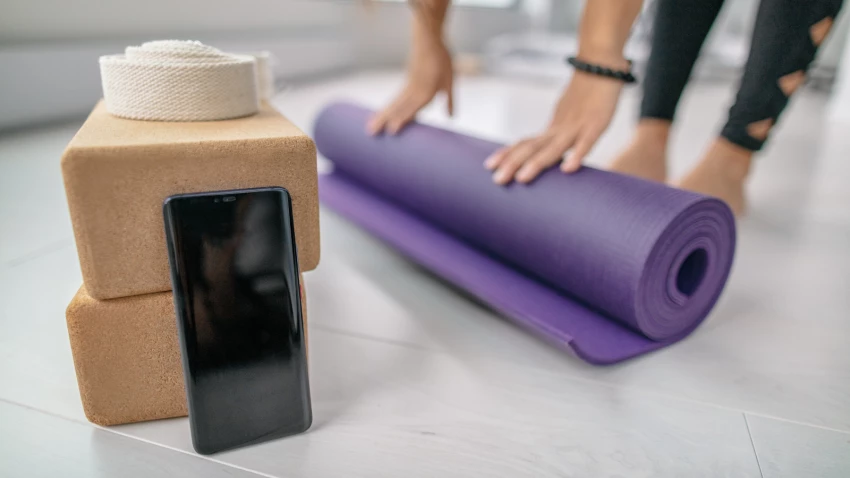 Student rolling out mat getting ready for yoga class with mobile smartphone close by