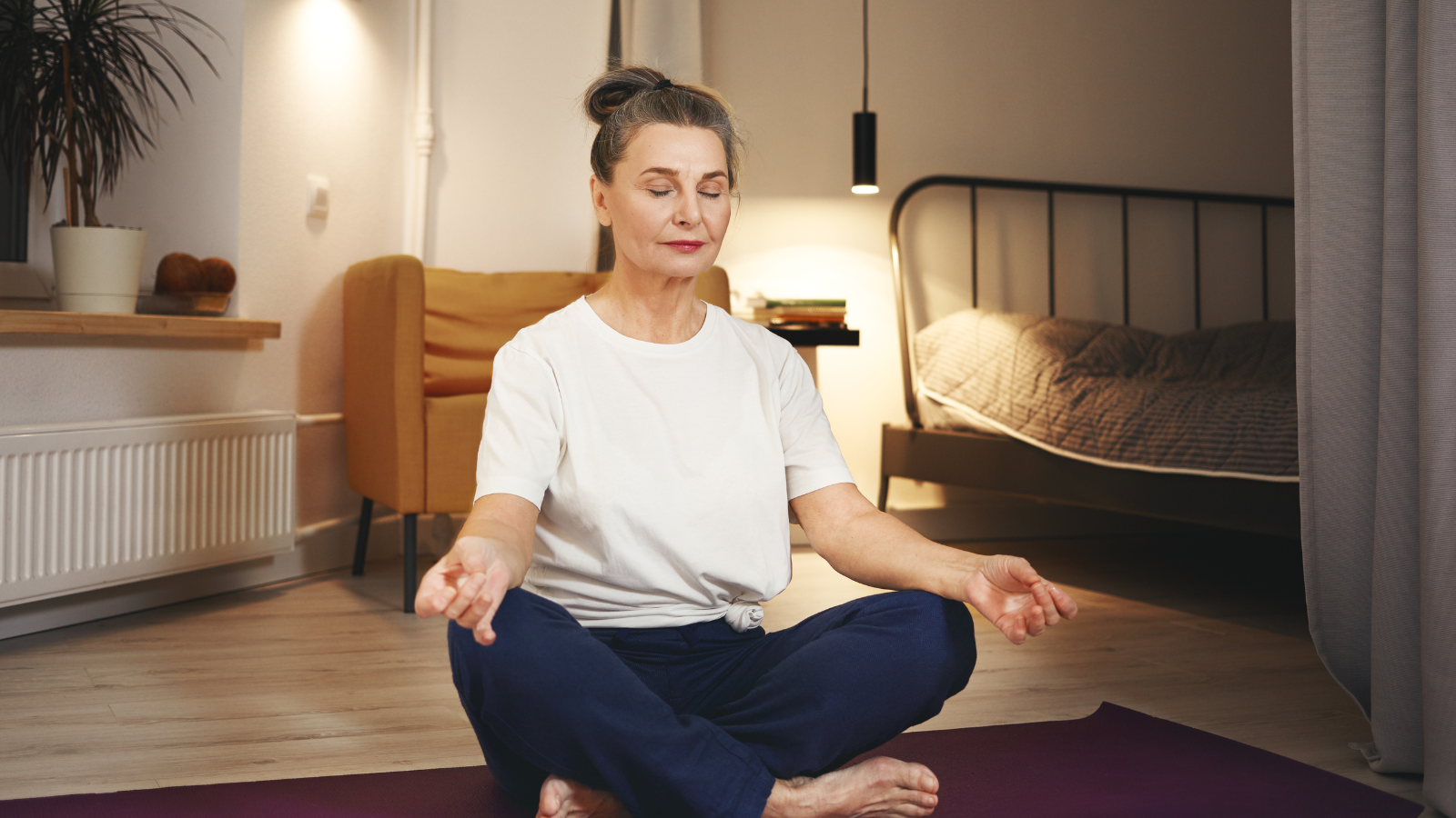 Senior woman in sportswear sitting barefooted on floor at home, keeping legs crossed on mat, doing meditation after yoga practice. 