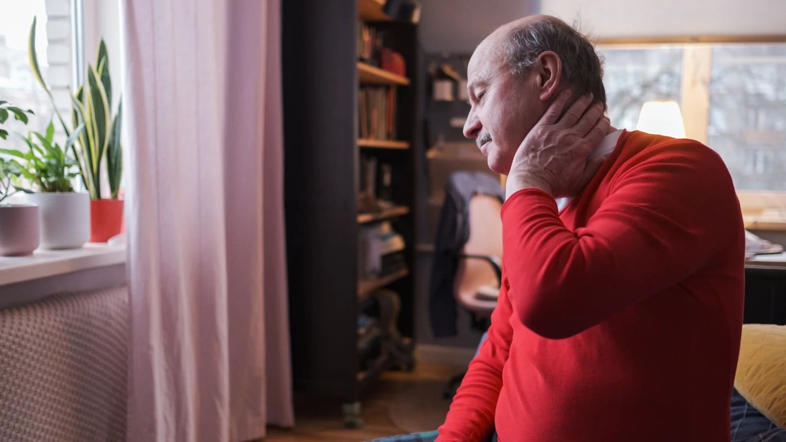 Frustrated senior man in red sweater holding hand on his neck sitting on sofa