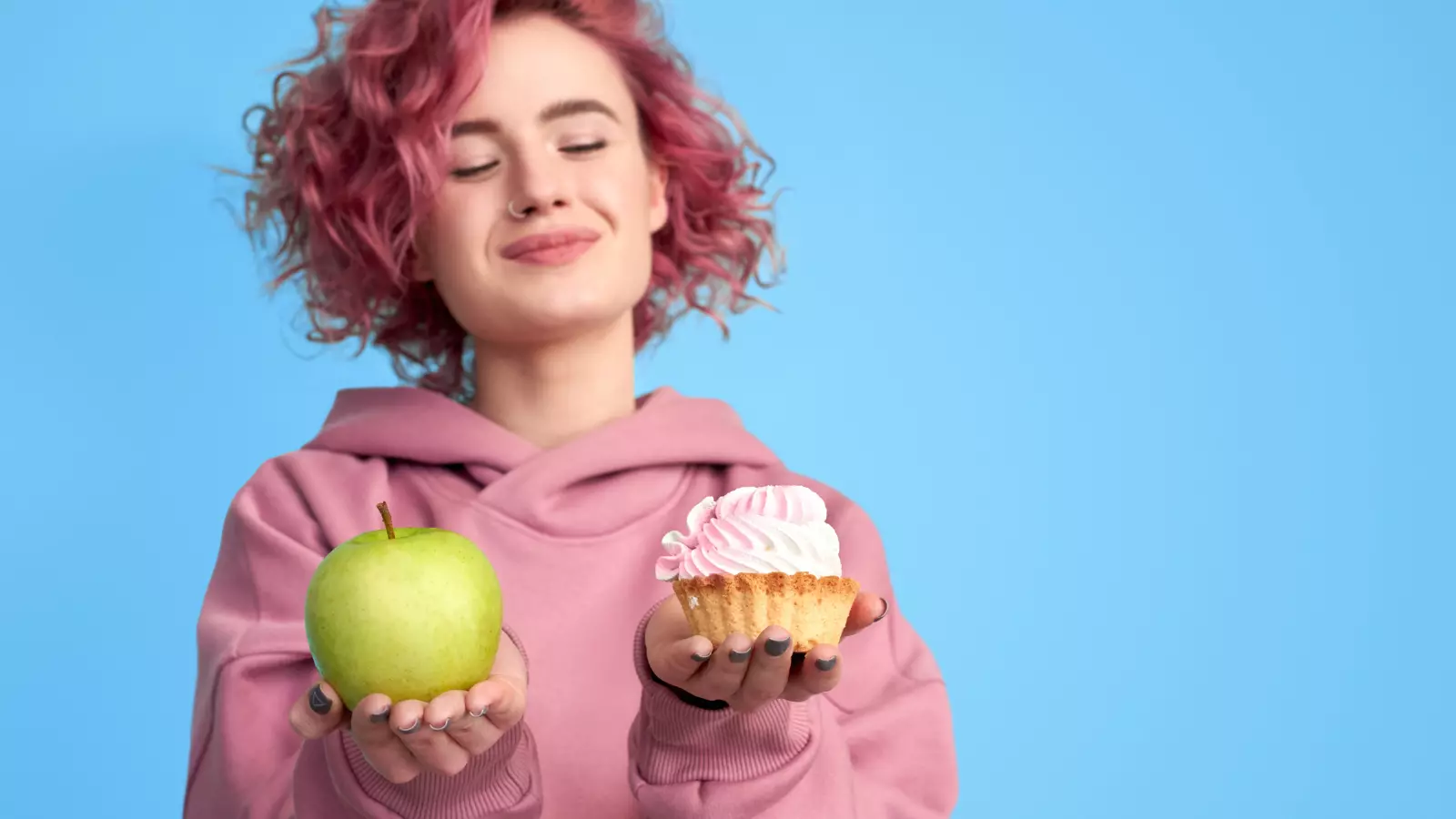 Pretty smiling girl with pink hair in hoodie comparing between fresh green apple and sweet cupcake in hands over blue background.