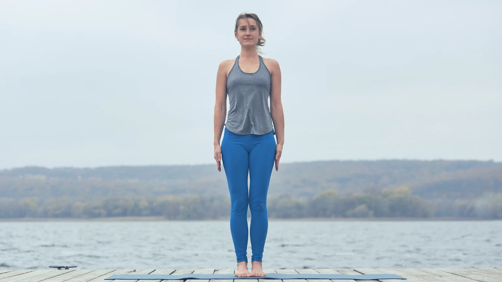 Beautiful young woman practices yoga asana Tadasana - Mountain Pose on the wooden deck near the lake