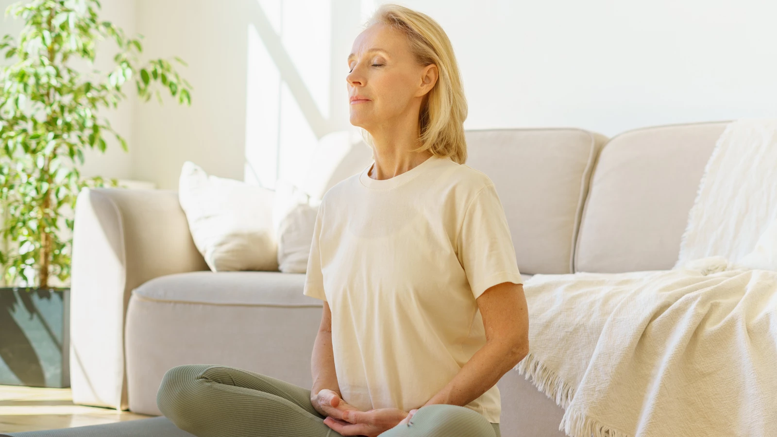 senior woman in lotus position meditating with closed eyes at home while sitting on yoga mat