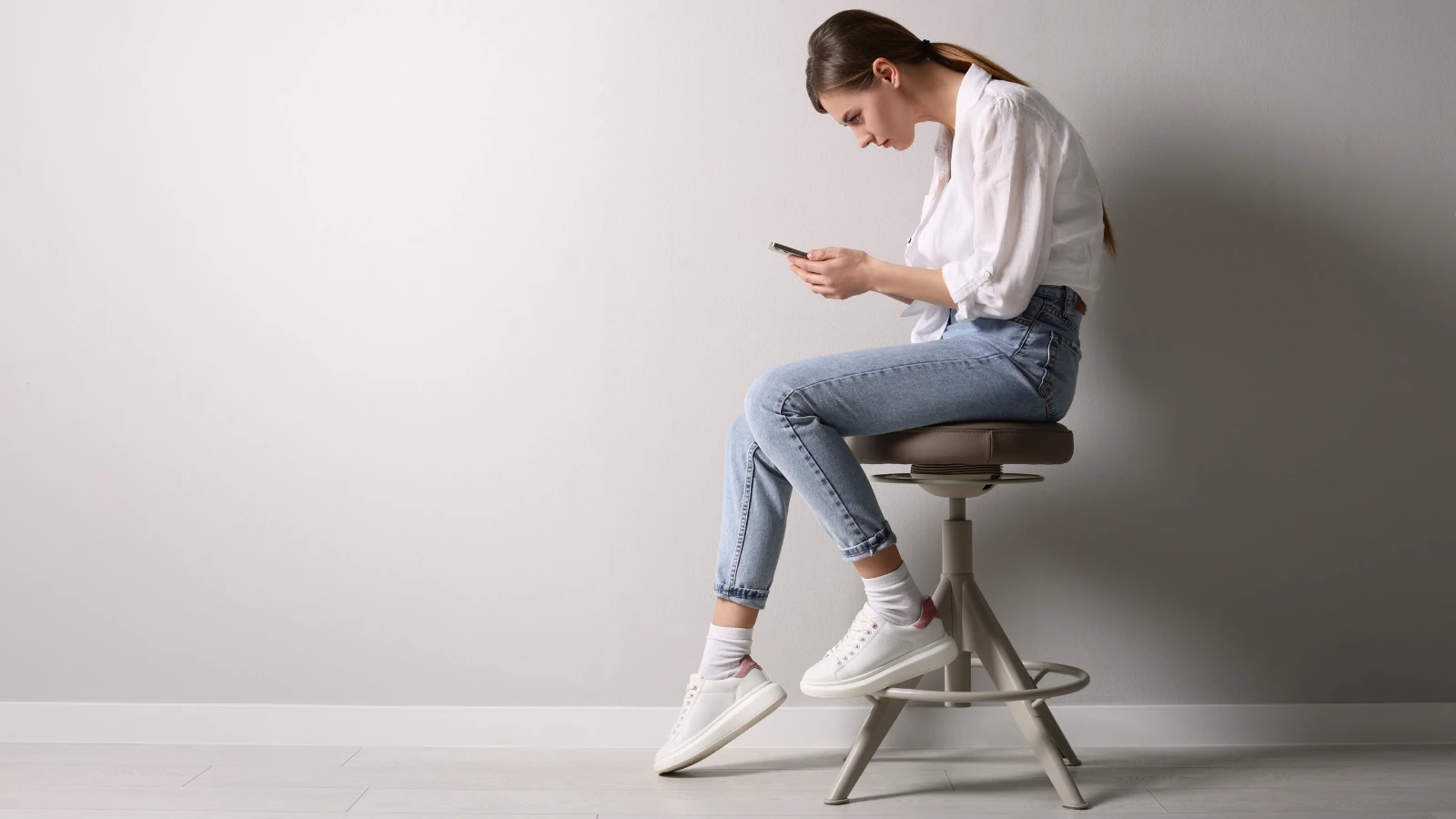 Woman with bad posture using smartphone while sitting on stool near light grey wall.