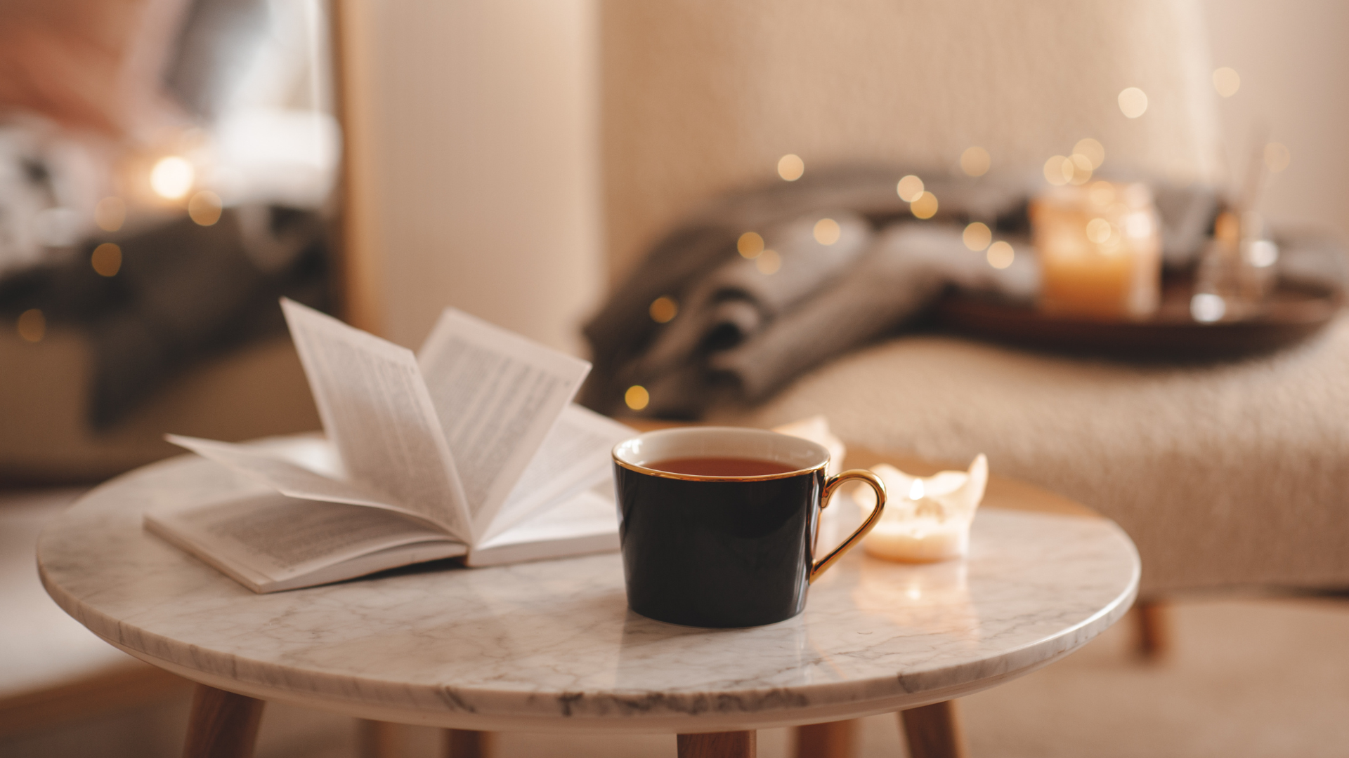 Cup of tea with paper open book and burning scented candles on marble table over cozy chair and glowing lights in bedroom closeup. Winter holiday season. 