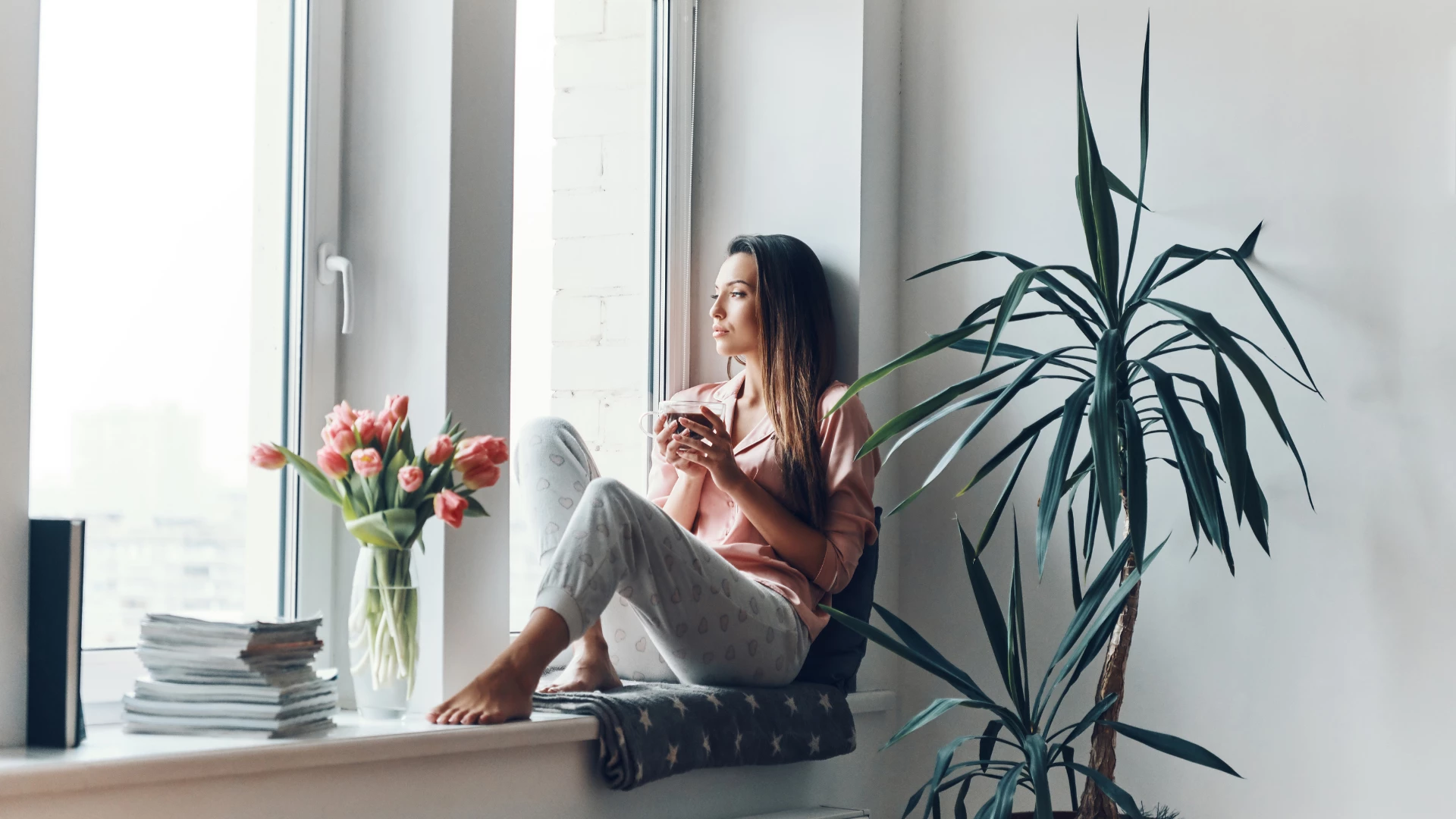 Thoughtful young woman in cozy pajamas looking through the window while resting at home