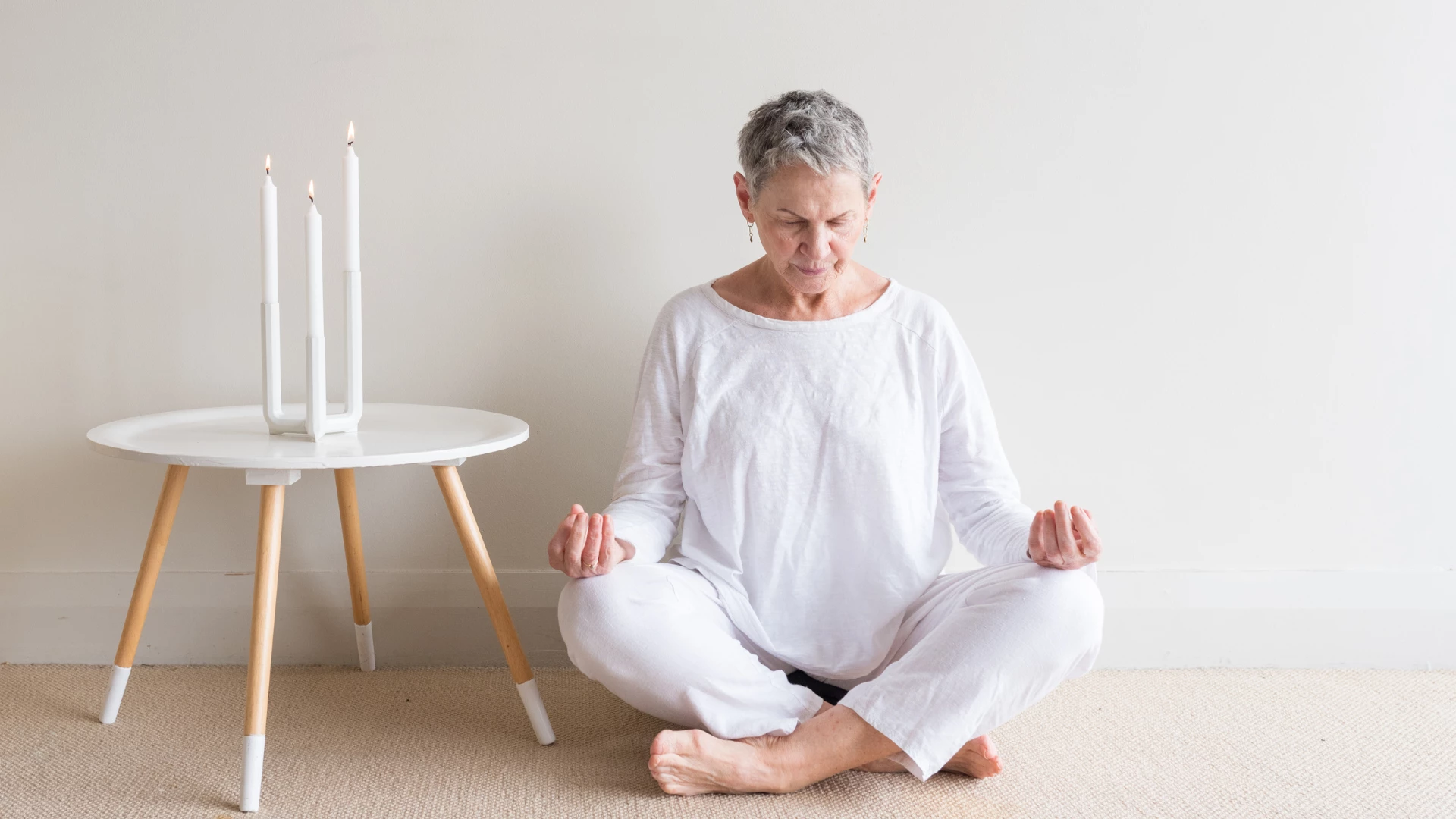 Beautiful older woman in white clothing sitting in yoga meditation position with candles against neutral wall background