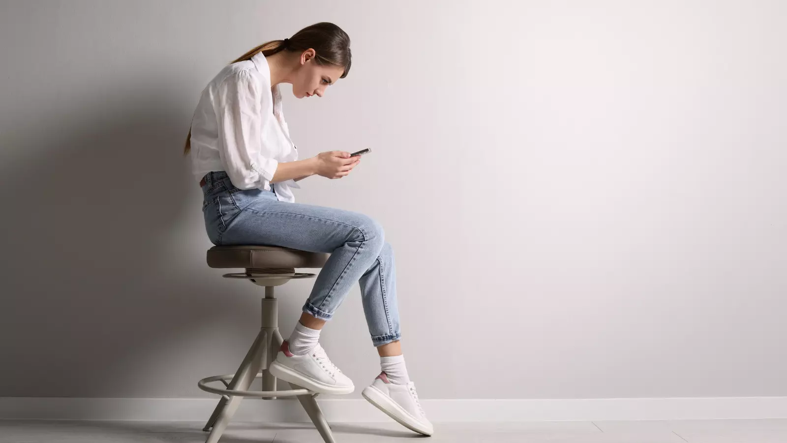 Woman with bad posture and text neck using smartphone while sitting on stool.