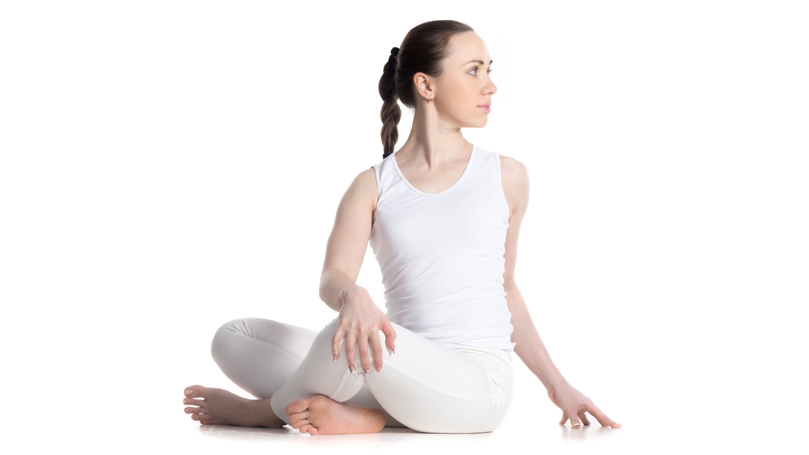 Sporty beautiful young woman in white sportswear practicing yoga, sitting cross legged in Revolved Easy Pose, a spinal twist also known as Parivrtta Sukhasana, while maintaining healthy posture.