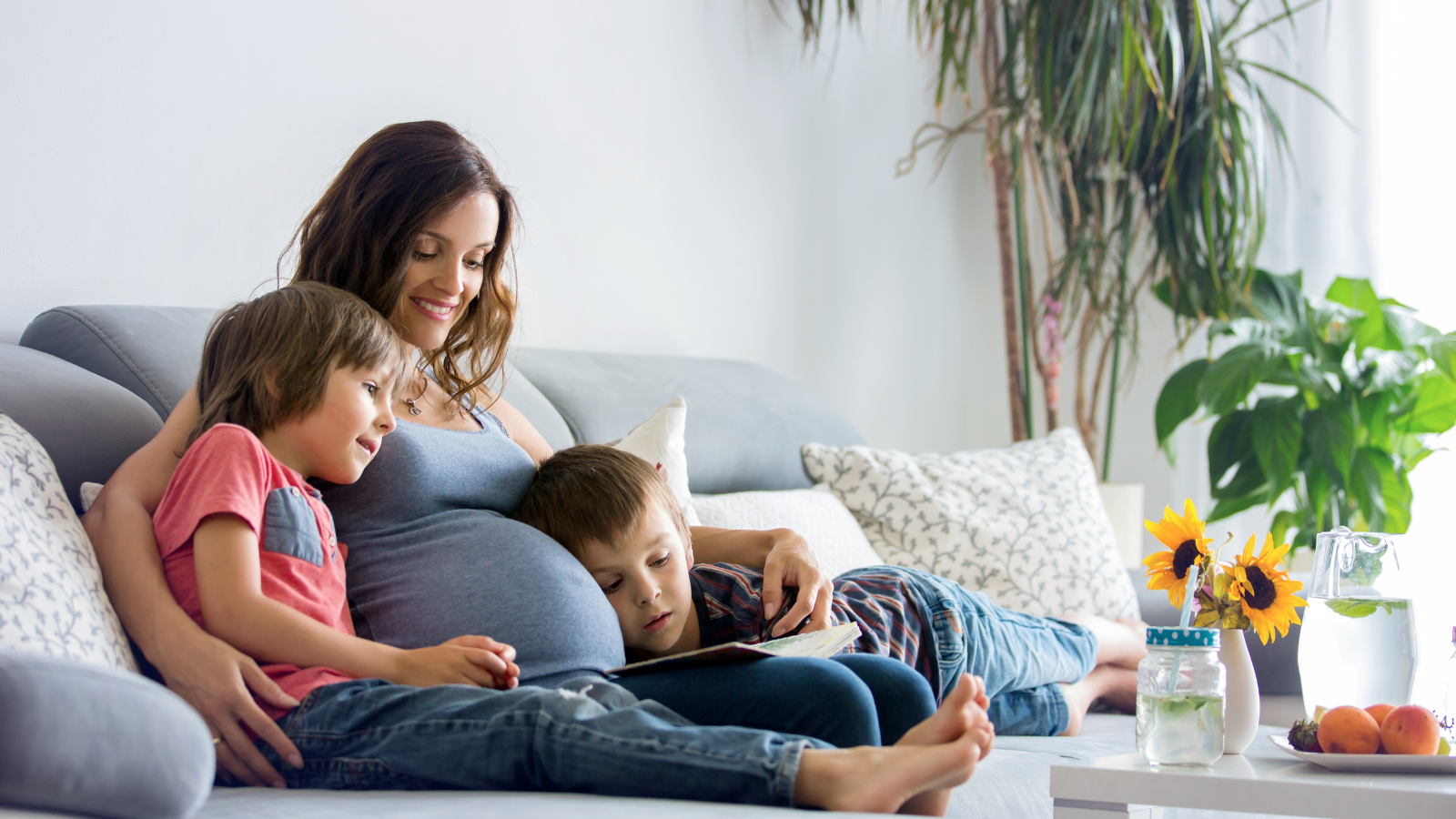 Young pregnant woman, reading a book at home to her two boys, eating fruits, hugging and laughing