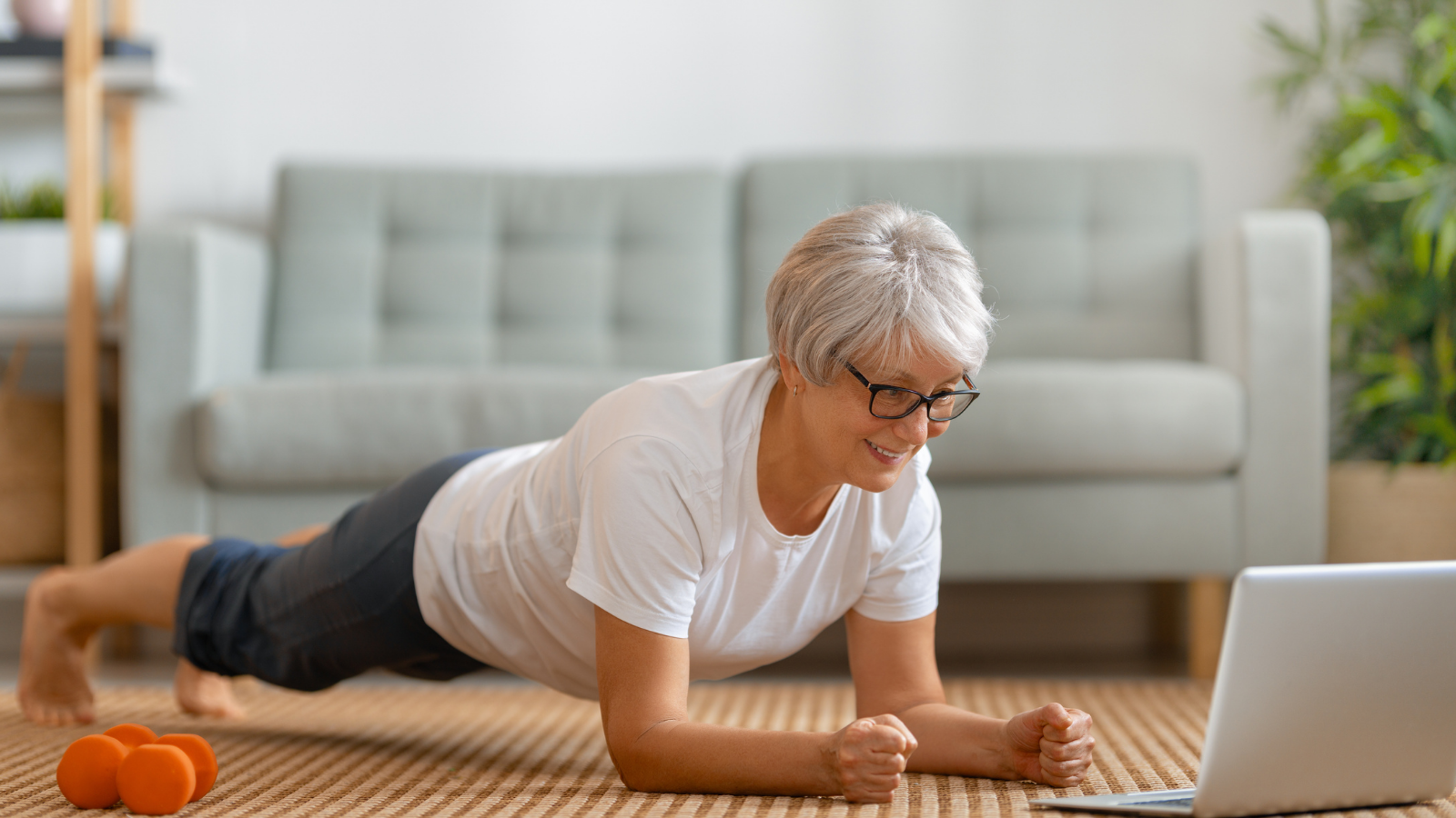 Senior woman in front of laptop in Plank Pose (Phalakasana) while participating in online yoga teacher training.