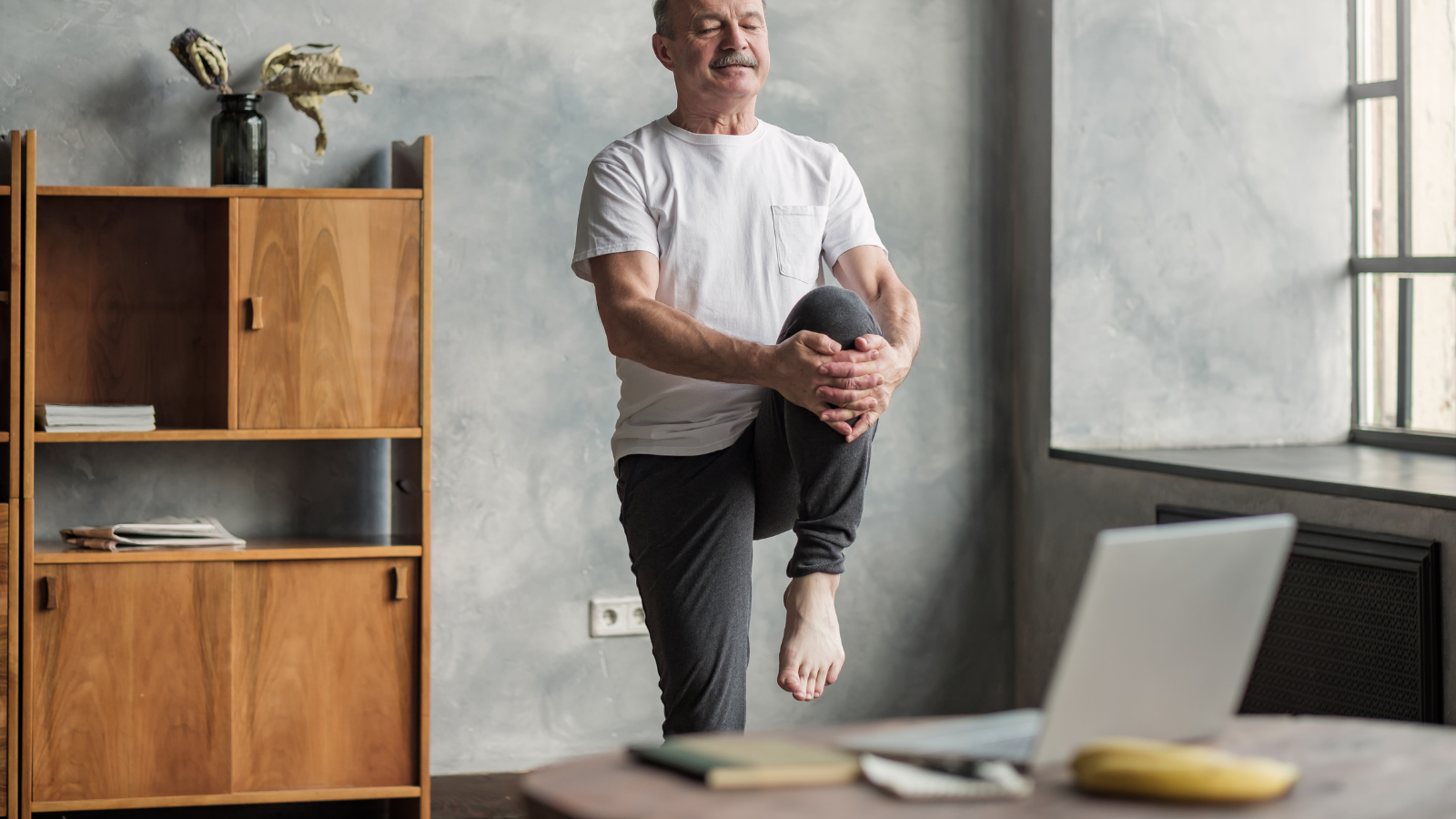 Senior man in front of laptop in Standing Knee to Chest Pose (Tadasana Pawanmuktasana), participating in an online yoga teacher training.