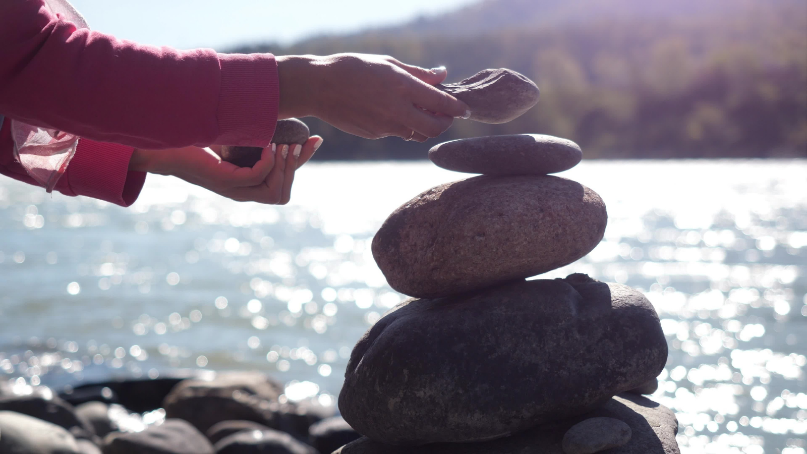 A woman stacking pebbles on the shore next to a river. 