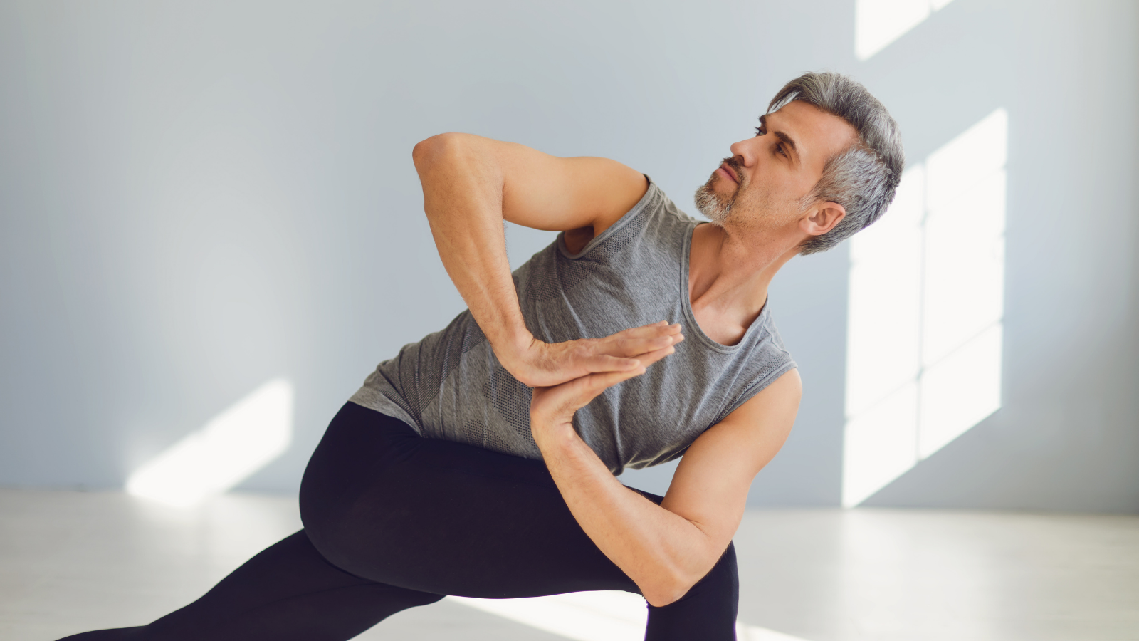 A mature man in sportswear is practicing yoga balance on the floor in a gray room