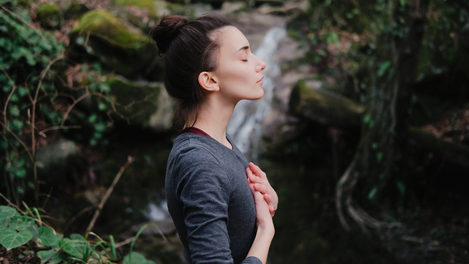 Young woman practicing breathing yoga pranayama outdoors in moss forest on background of waterfall. Unity with nature concept