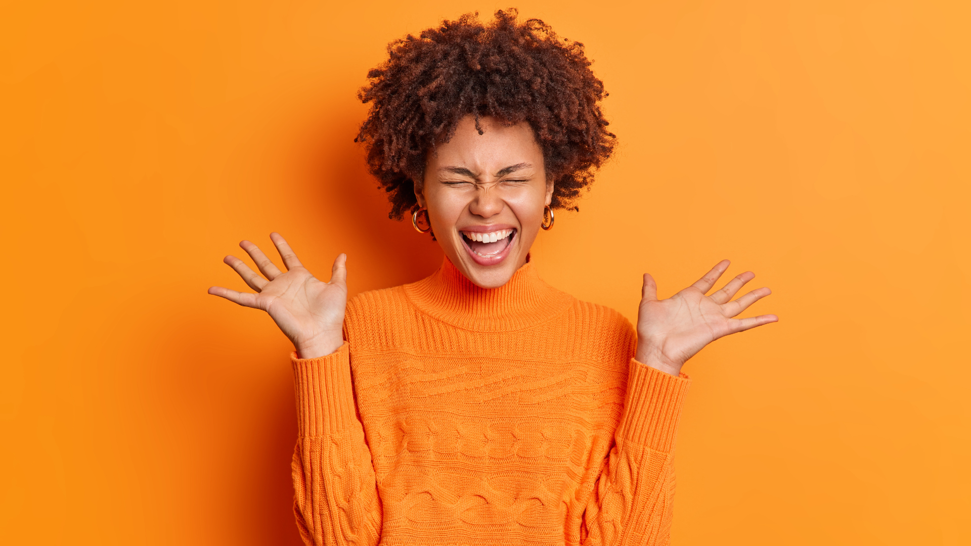  Positive overjoyed young African American woman raises palms feels very glad expresses joy dressed in casual jumper isolated over orange background. 