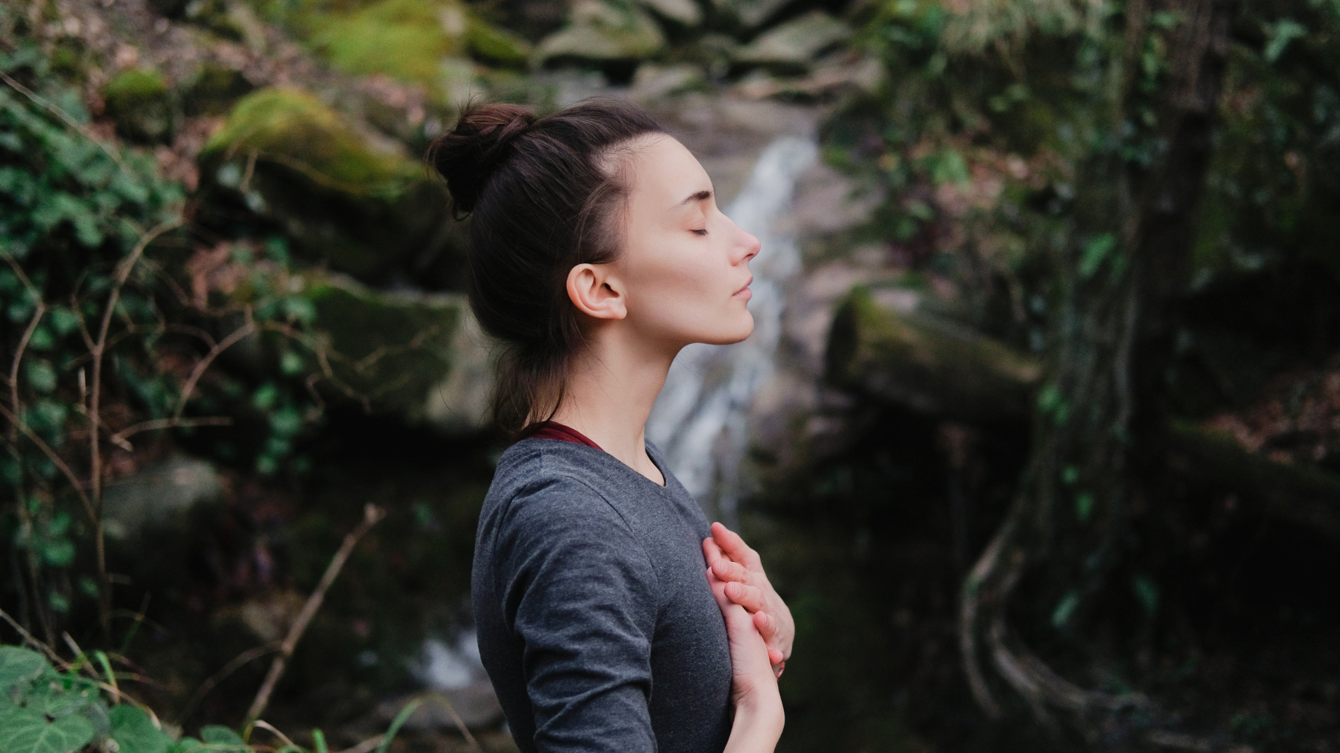 Young woman practicing breathing or yoga pranayama outdoors in moss forest on background of waterfall. Unity with nature and contentment concept.