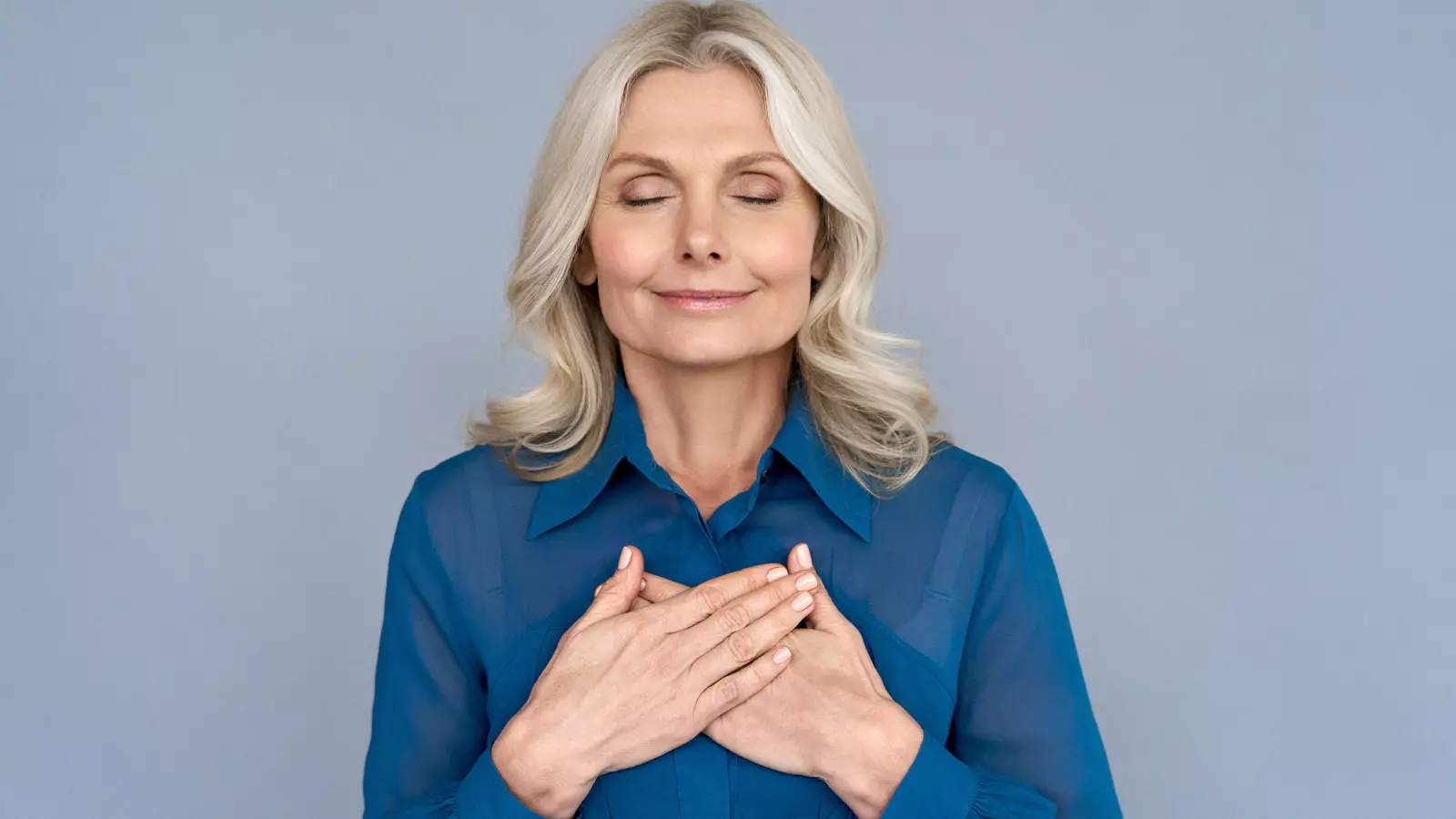 Happy mindful thankful middle aged old woman holding hands on chest meditating with eyes closed in gratitude, mental health balance, peace of mind concept, heart health concept.