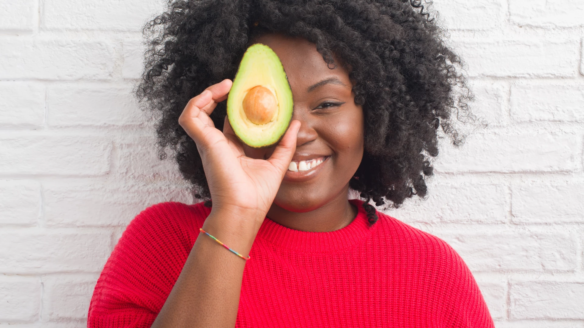 Young African-American woman over white brick wall eating avocado as a healthy part of a yoga diet.