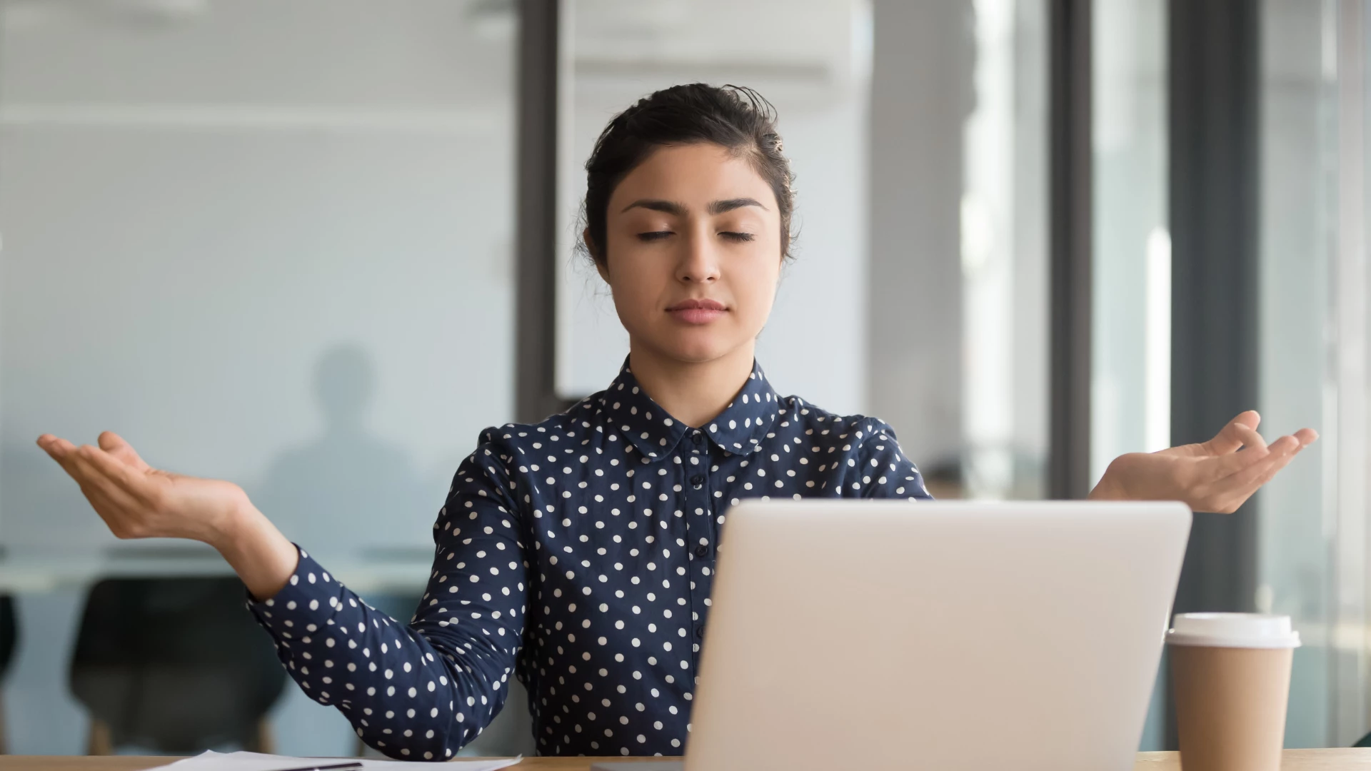 Calm office worker Hindu woman sitting at her desk near laptop with fingers in mudra gesture. Resting at workplace with yoga to relieve neck and eye tension.