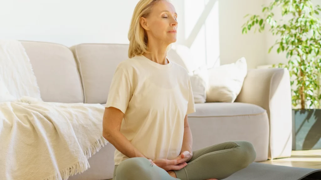 Peaceful and calm senior woman in Lotus Pose meditation and her breathing practices with closed eyes at home.