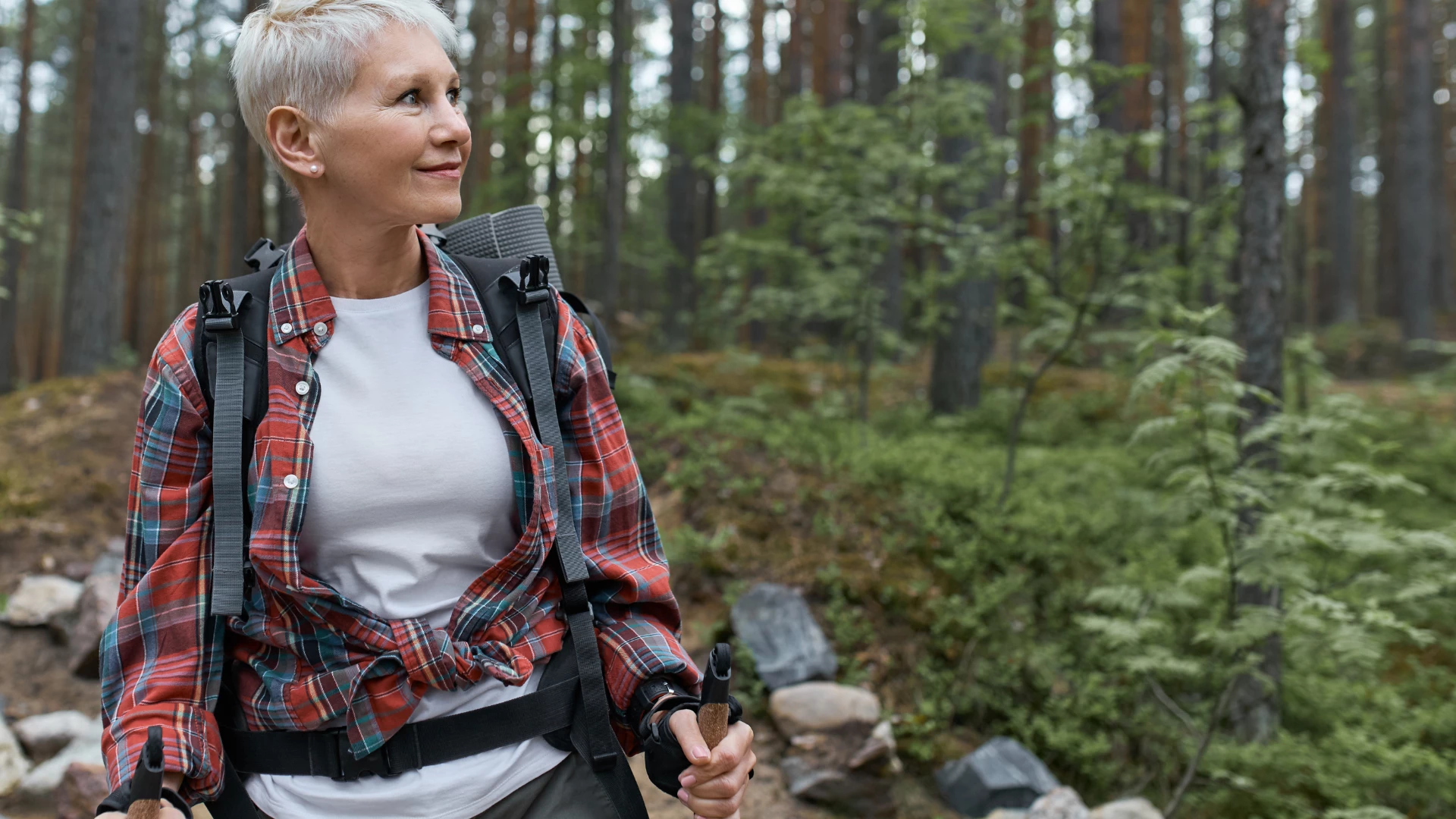 Outdoor portrait of happy European female pensioner with backpack and poles, enjoying beautiful nature while Nordic walking in pine forest. Aging, people, active lifestyle and health lungs concept, Lung elasticity.