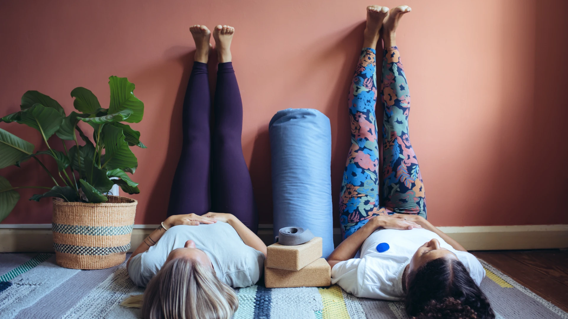 Two women in legs-up-the-wall yoga pose with props, illustrating relaxation benefits and the link between yoga and gene expression for stress